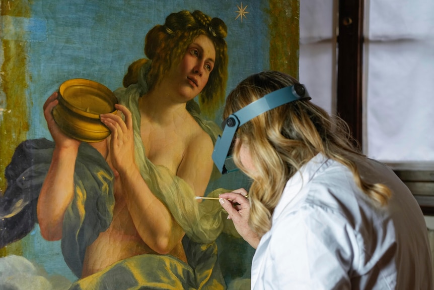 a woman in white coat uses a small brush on the baroque nude of a woman