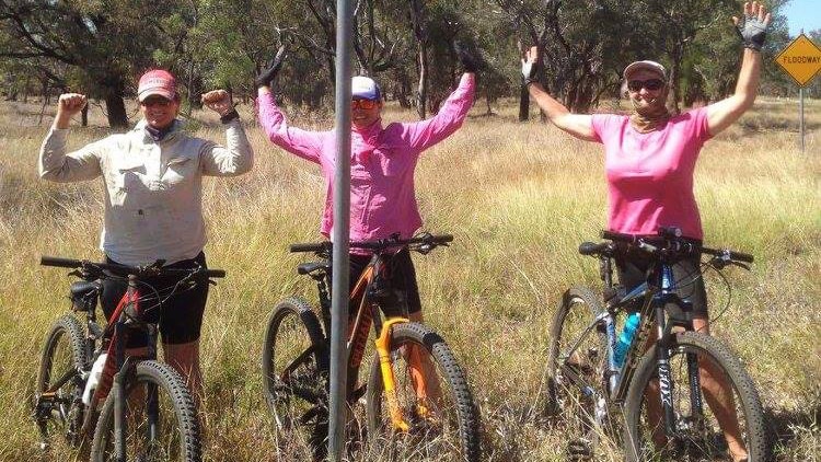 Three women stand with their mountain bikes, arms in the air, underneath a sign that reads 'Belyando River'