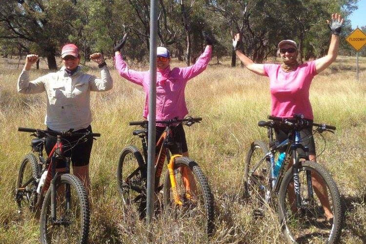 Three women stand with their mountain bikes, arms in the air, underneath a sign that reads 'Belyando River'