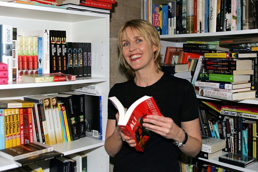 A smiling woman stands between packed bookshelves, holding an open book.