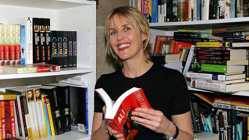 A smiling woman stands between packed bookshelves, holding an open book.