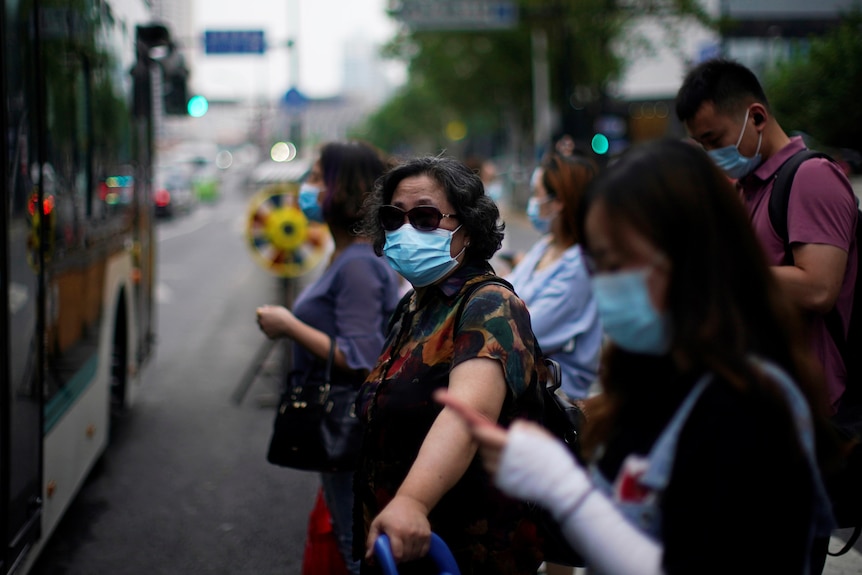 A woman wearing a surgical mask and dark sunglasses looks at the camera, standing in a crowd waiting for a bus