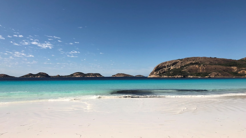 Empty beach with clean white sand and clear water, rocky island in the sea in the background. 