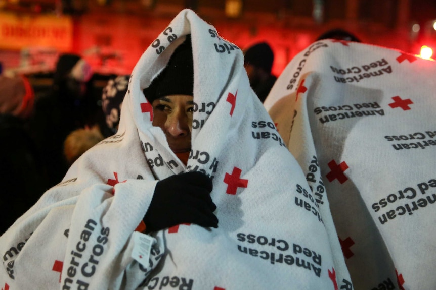 A woman wearing a beanie and gloves wraps her head and shoulders in a red cross blanket after evacuating from apartment fire.