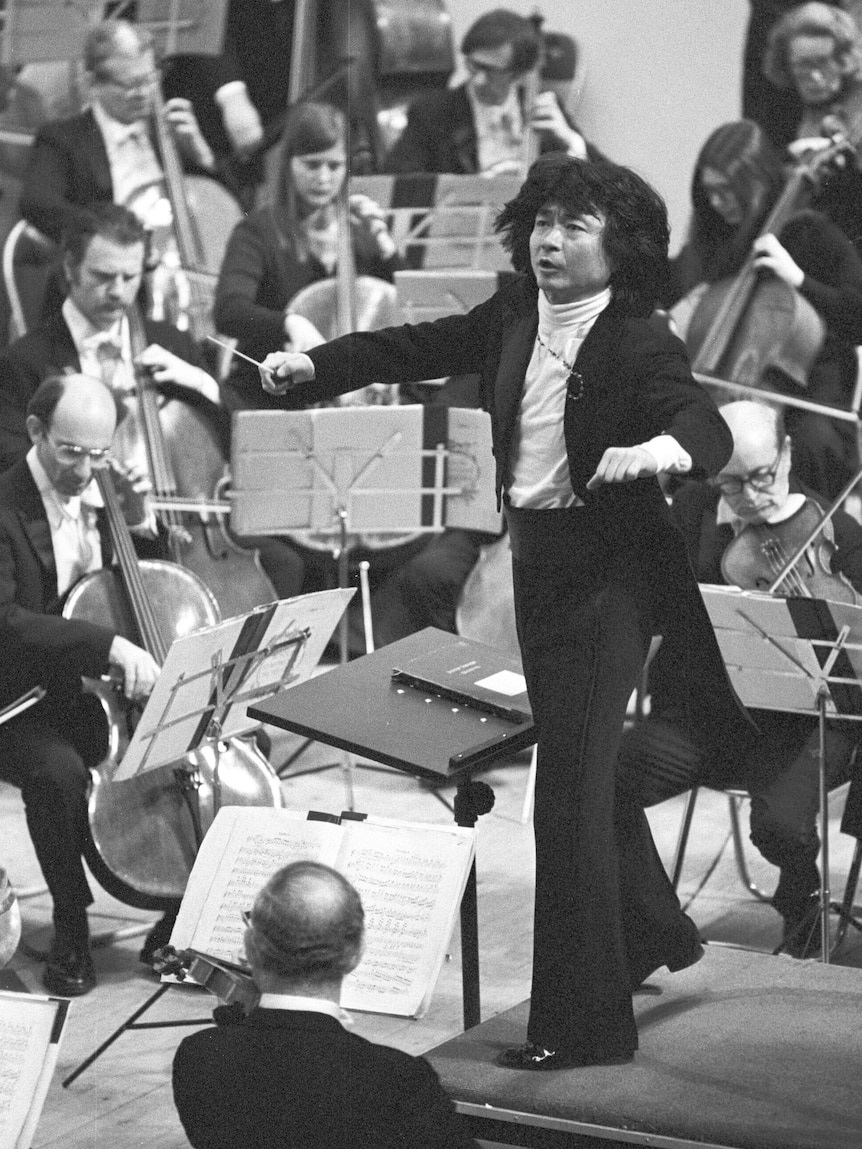 A black and white photo showing a young man wearing a black suit raising his arms to conduct an orchestra