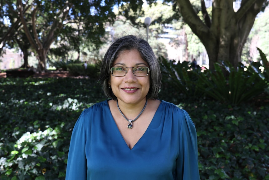 A photo of Dr Sureka Goringe wearing a blue top, and glasses. Behind her is a nature backdrop with trees and bushes.