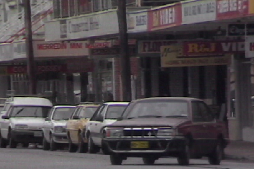 A 90s/2000s car in a street pulls out onto the road, with other sedans parallel parked behind it. A sign sign reads 'Merredin'.