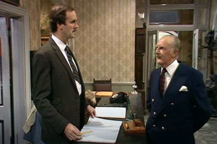 Cleese and Berkeley stand speaking at the reception desk
