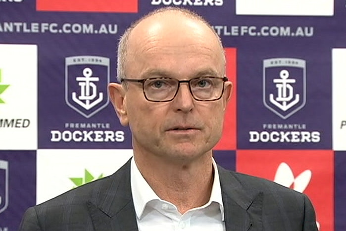 A headshot of Dale Alcock speaking in front of a board of sponsor logos.