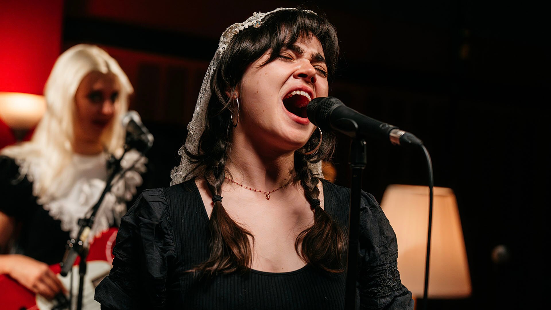 Person with brown hair in plaits, eyes closed singing into a microphone. A person with blonde hair in the background