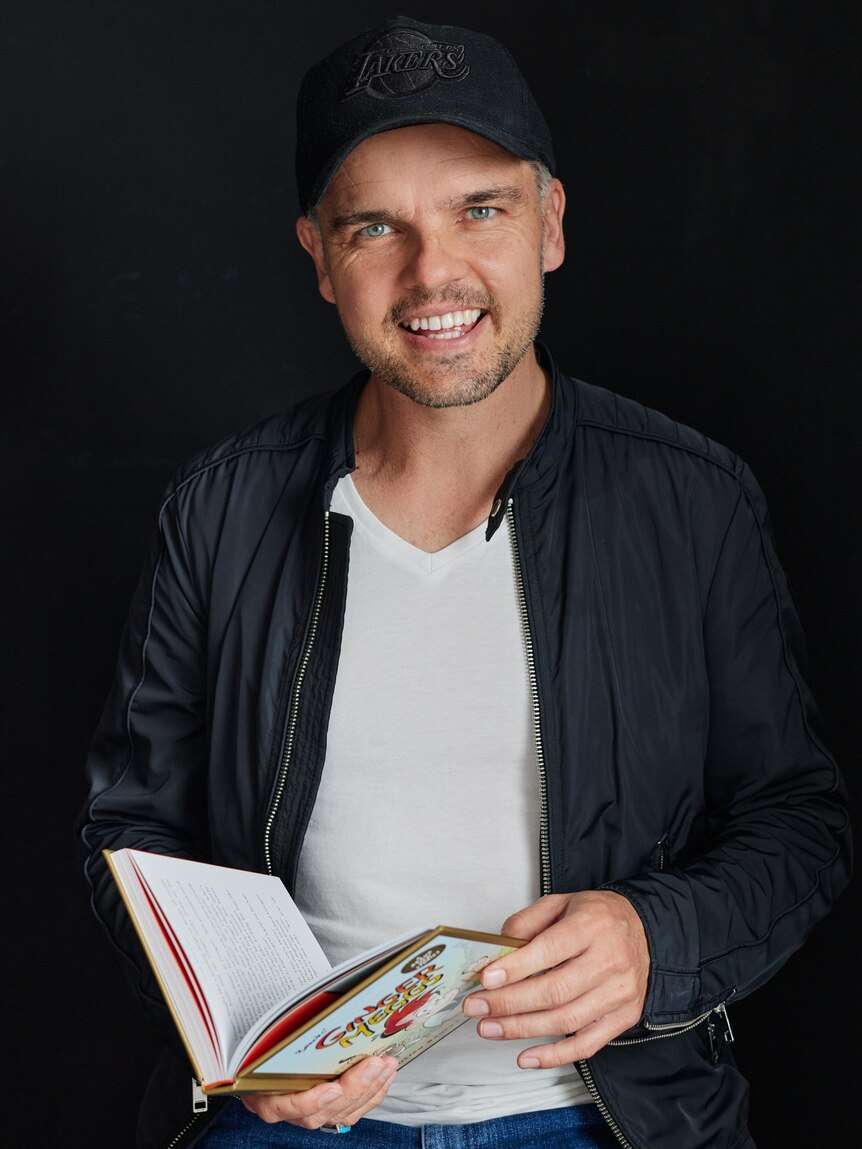 A man wearing a black hat and jacket holds open a children's book and smiles.