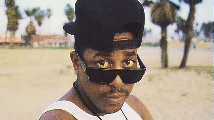 Tone Loc wears a white singlet, backwards baseball cap and sunglasses. He is at the beach posing for the camera.