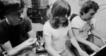 AC/DC producer George Young sitting at a piano with brothers Malcolm and Angus Young.