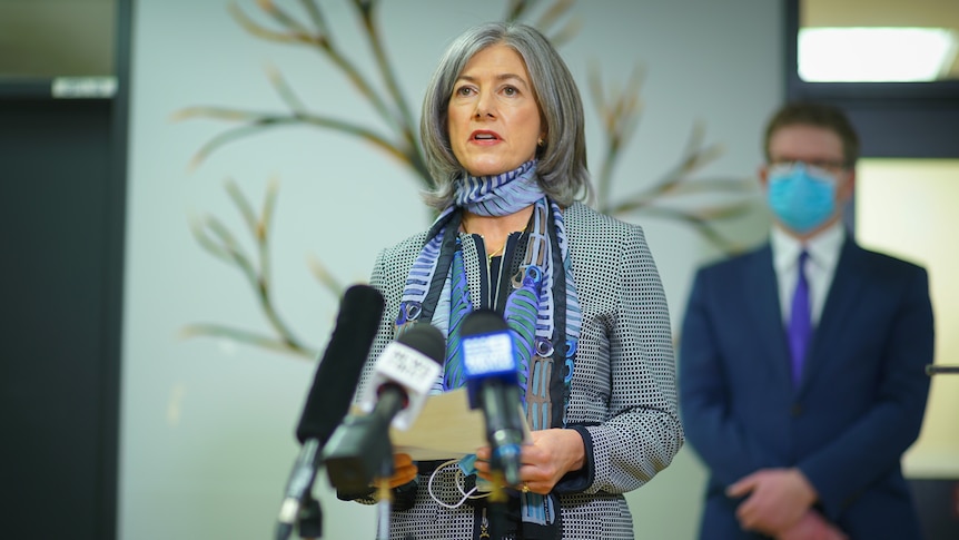 A woman with grey hair wearing a checked jacket and a purple scarf with a man standing behind her