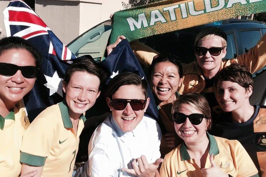 A group of women in green and gold shirts smile by a van, holding up a Matildas banner.