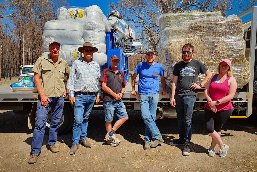 A group of people stand in front of a truck loaded with hay and bags of stock feed.