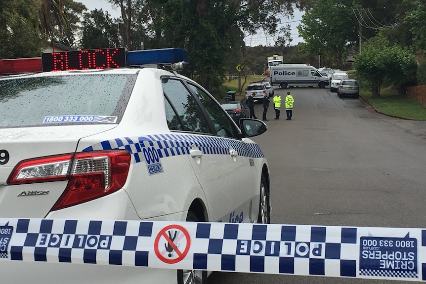 A police car in the foreground in a Davidson street as officers stand outside of a crime scene where four people were found dead