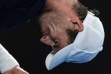 A man holds a tennis racquet in both hands and grimaces, bent over double with his eyes shut