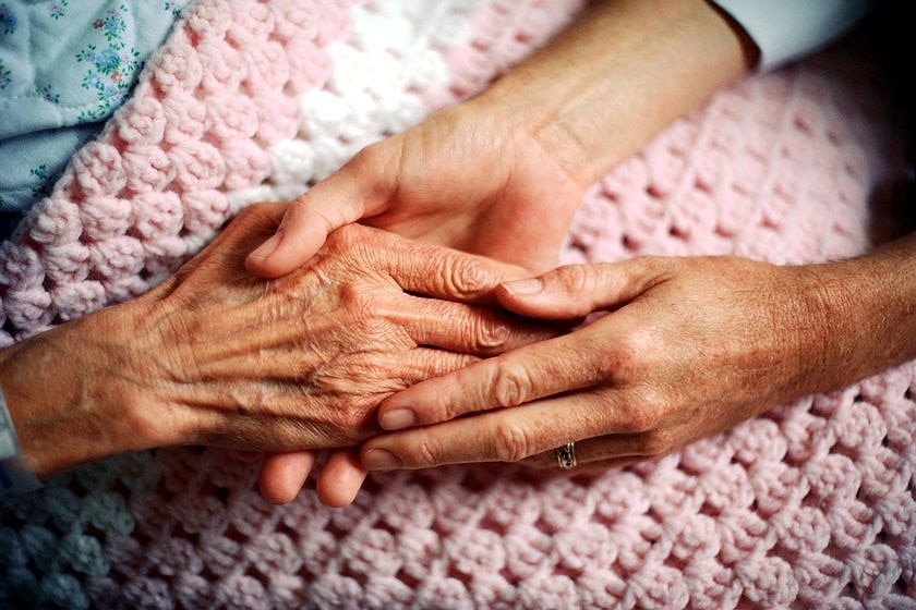 An elderly woman's right hand is held by both hands of a younger person on top of a pink throw with a white stripe