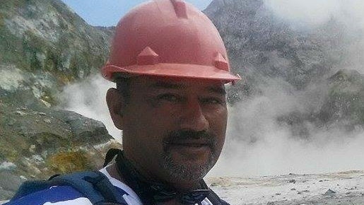 A man in a hard hat stands in front of a scene of volcanic rock, with steam in the background.