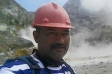 A man in a hard hat stands in front of a scene of volcanic rock, with steam in the background.