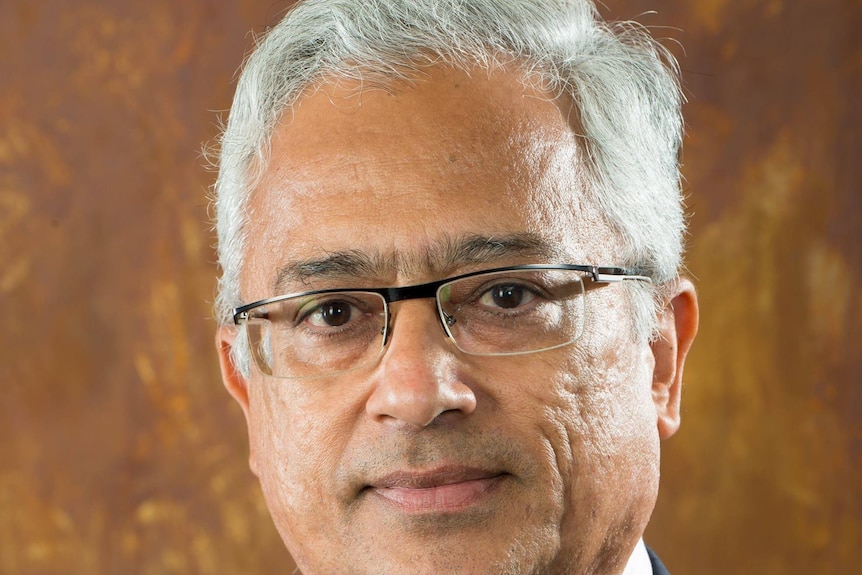 A middle-aged Indian man with white hair dressed in dark suit, white shirt and pink tie looks at camera.