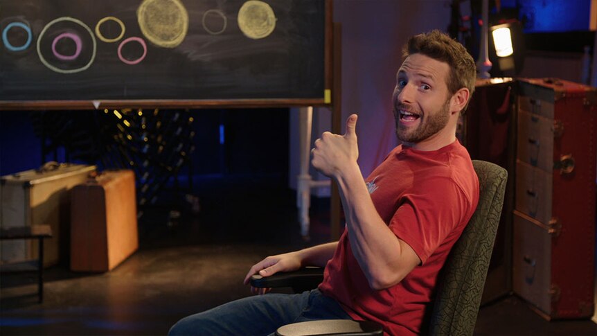 A smiling man sits with thumbs up in front of a blackboard with circular shapes