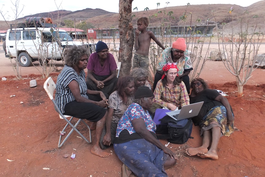 Members of an Aboriginal community crowd around a woman using a laptop.