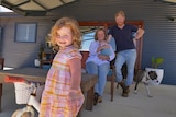 A smiling toddler stands on a deck in front of her beaming parents. Her mum hold a baby.