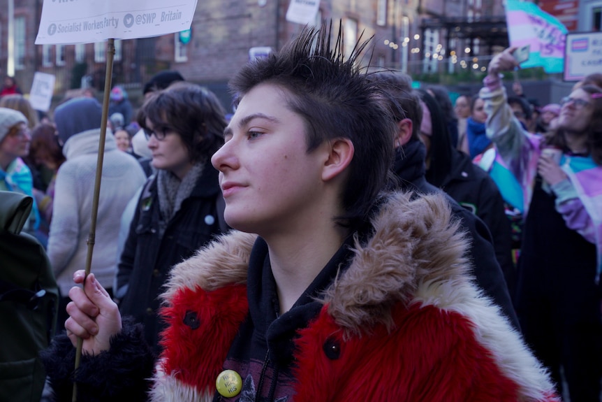 A person with short, spiked brown hair stands amongst a crowd holding a sign out of frame
