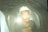 A man trapped inside a tunnel wears a white hard hat as he looks into a camera for a low resolution picture.
