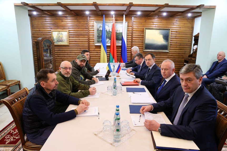 Four officials sit in a row along a table opposite five suited officials, with three flags in the background of the room. 