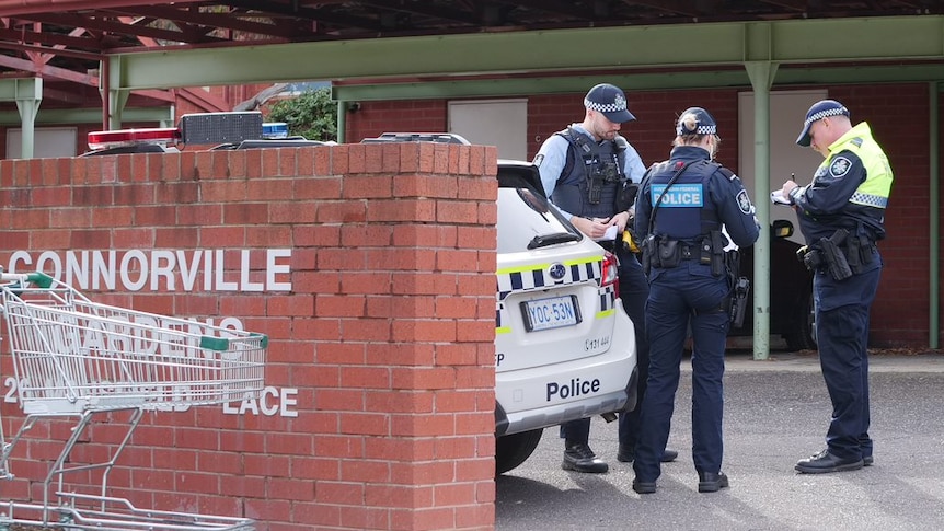 Police stand in the parking area of a unit complex, with trolleys strewn in the garden.