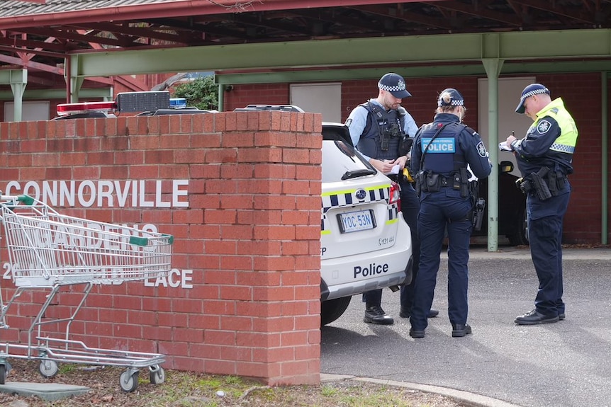 Police stand in the parking area of a unit complex, with trolleys strewn in the garden.