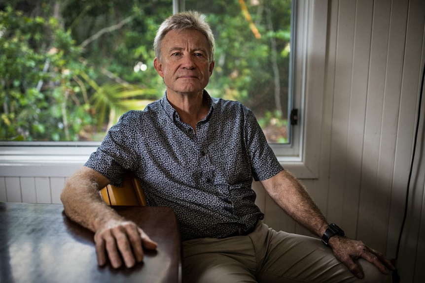 A man sits in a chair inside a home with greenery seen out a window behind him.