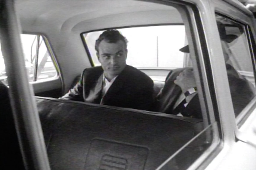 A historical black-and-white image of a criminal dressed in a suit in the back of an unmarked police vehicle