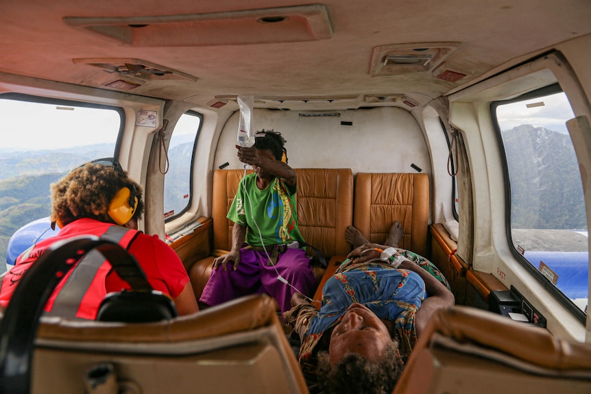 A women lies in a helicopter, while two others look over her. One holds her IV drip.