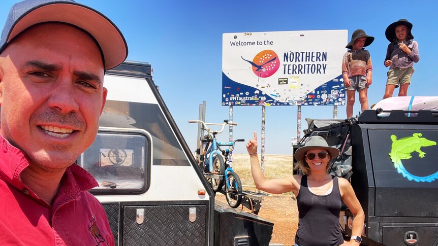 2 people stand near a black four-wheel-drive and a caravan, in front of a Welcome to the Northern Territory sign. 
