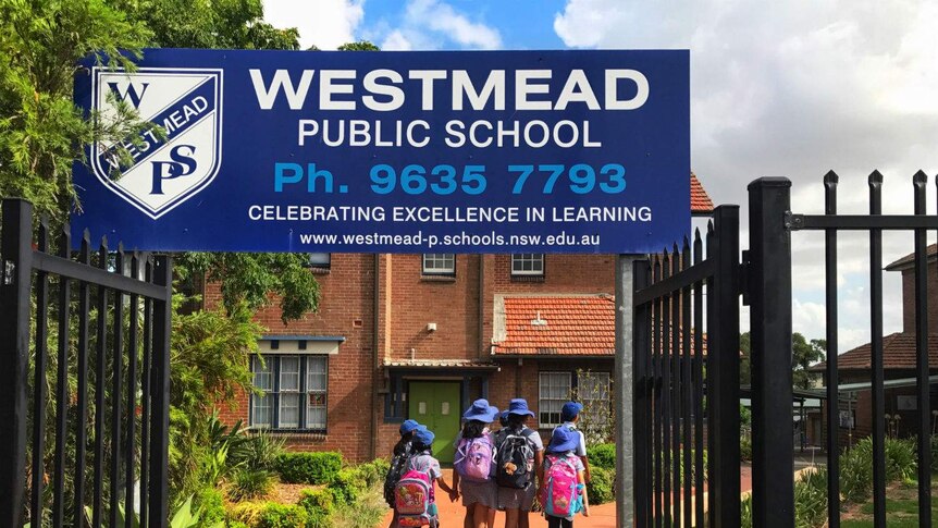 The Westmead Public School sign on top of a black fence with school children walking in the background.