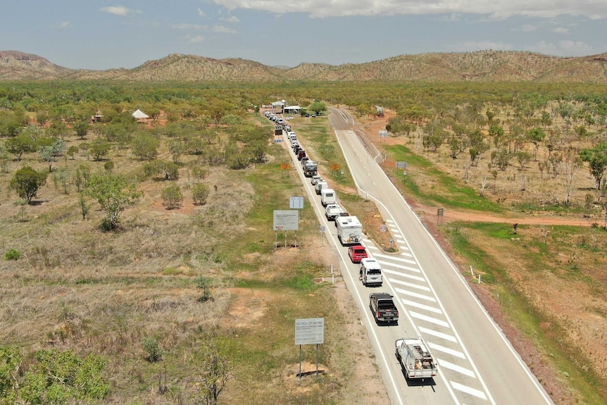 A line of vehicles at the WA–NT border, including cars, caravans, work utilities and small trucks