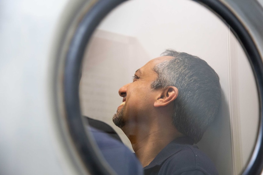 Looking through a round window into an airlock, a man is smiling while waiting to get into the secure lab on the other side.
