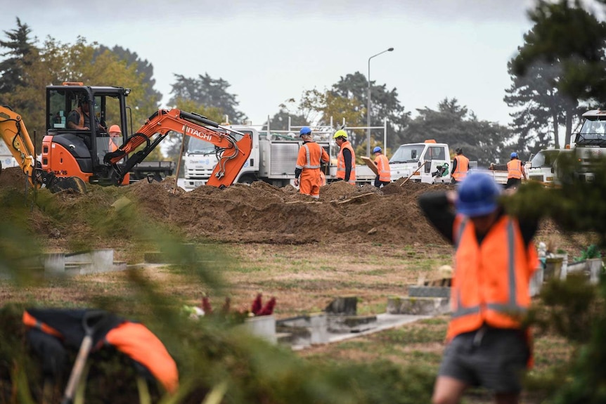 Workers are seen in orange hi-vis jackets as graves are prepared with excavation equipment seen in the background on a grey day.
