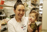 An woman in a white chef's uniform in a kitchen holding her six-month old baby