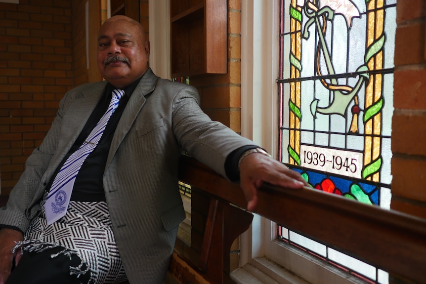 Reverend Maile Molitika sitting in front of glass stained window, wearing a grey jacket and traditional dress. 