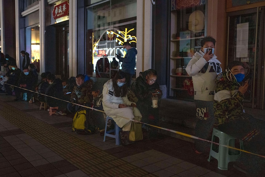 People line up while sitting on little seats outside a shopfront at night