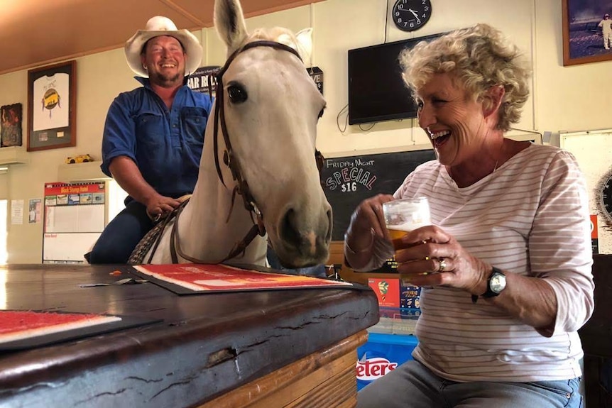 Woman sat at a pub bar with a beer laughing next to a horse and it's rider also standing at the bar inside pub. 