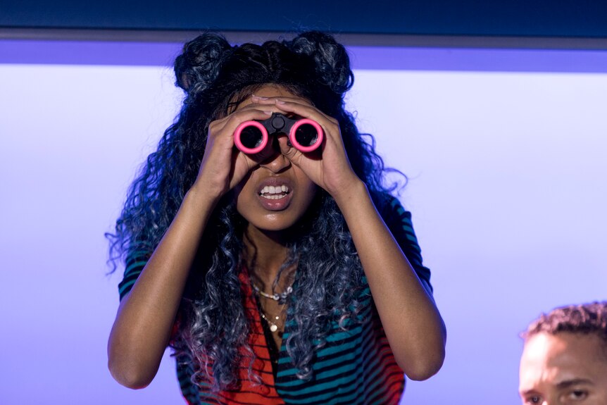 On stage, a girl with long hair squints through a pair of binoculars. The lenses are rimmed in hot pink.