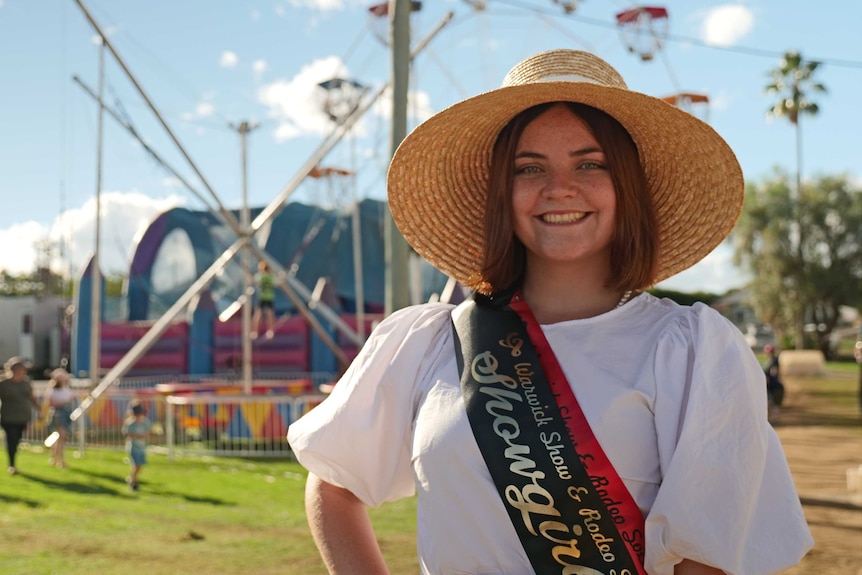 Smiling red-haired girl in a white shirt, showgirl ribbon sash and large broad-brimmed hat.