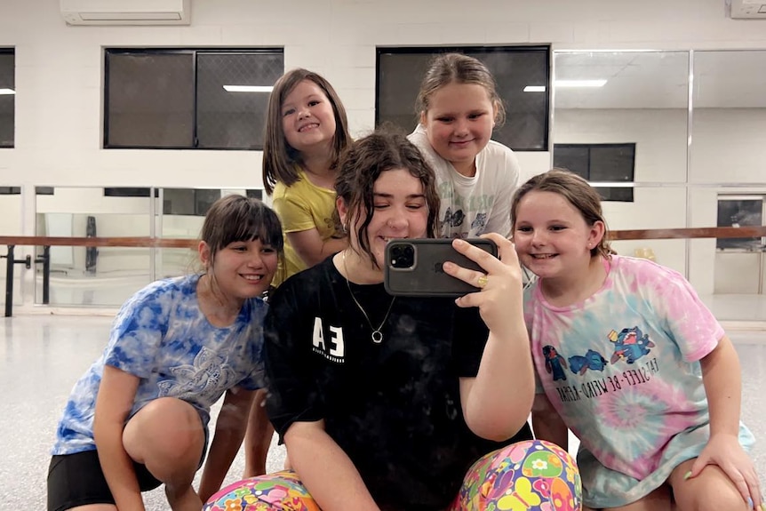 A young woman wearing a black t-shirt takes a selfie in a dance hall mirror surrounded by kids
