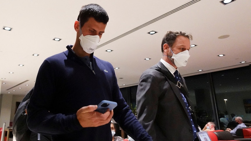 Two men walking, while wearing masks. man on the left looking down at his phone. 